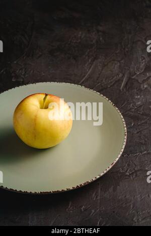 Fresh sweet single apple in grey plate on dark black textured background, angle view Stock Photo