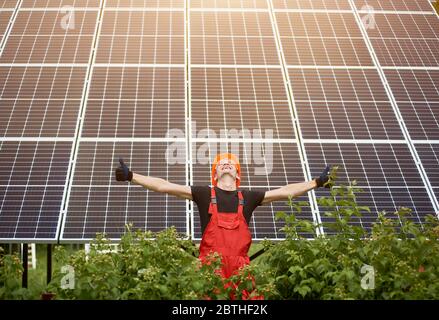 Happy worker raising his hands, showing thumbs signs like on a background of photovoltaic panels solar station. Man in orange uniform. Science solar energy. Renewable energy. Stock Photo