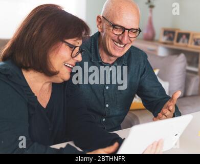 Senior middle aged happy couple embracing using tablet together -  smiling elderly family reading the news and shopping online from home - mature peop Stock Photo