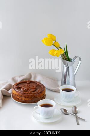 Homemade cake, two cups of coffee and tulips in a vase against a light background. Home baking concept. Vertical frame. Stock Photo