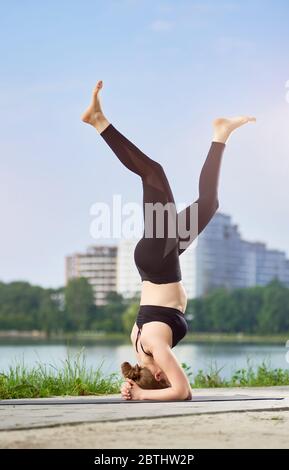 Yoga girl doing asana Shirshasana, standing on her head in the background of urban landscape under a blue sky. Woman working out in black pants and bra. Morning training outdoors at the summer Stock Photo