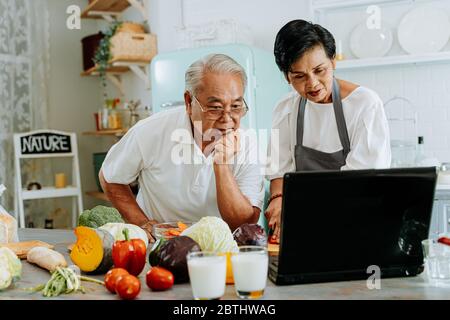 Senior Asian couple using a laptop while cooking in kitchen at home. 70s Elderly man and woman in relationship retirement lifestyle concept Stock Photo