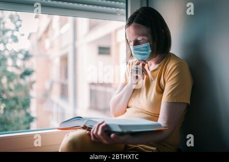 Woman in self-isolation reading book by the window. Female person with face protective respiratory mask in home quarantine.