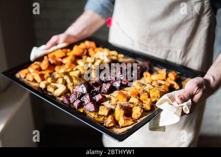 Male hands holding oven tray with baked vegetables mix. Vegetarian man cooking sweet potatoes and other veggies at home.