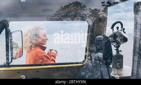 Young blond woman standing next to camper van overlooking the frozen winter sea. View through the open door. Stock Photo