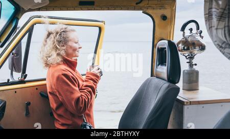 Young blond woman standing next to camper van overlooking the frozen winter sea. View through the open door. Stock Photo