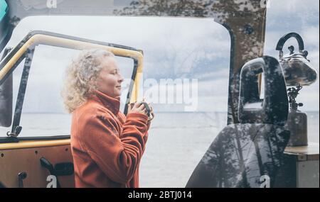 Young blond woman standing next to camper van overlooking the frozen winter sea. View through the open door. Stock Photo