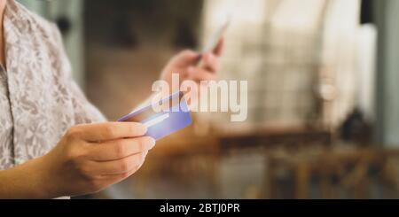 Cropped image of customer's hand holding a smartphone and credit card for doing a payment by using a credit card over blurred restaurant as background Stock Photo