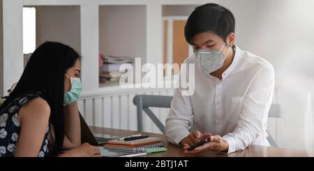 Photo of young couple that wearing a medical mask sitting and relaxing together at the wooden working desk over comfortable living room as background. Stock Photo