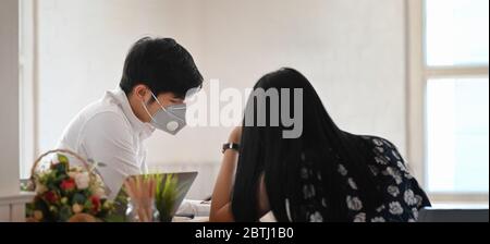 Photo of young couple that wearing a medical mask sitting and relaxing together at the wooden working desk over comfortable living room as background. Stock Photo