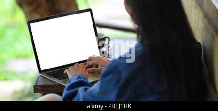 Woman typing on white blank screen computer laptop that putting on wooden working desk while sitting outside the house over nature as background. Stock Photo