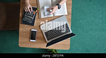 Above shot woman calculating and holding an Invoice while sitting at the wooden short legs table that surrounded by laptop and equipment over comforta Stock Photo