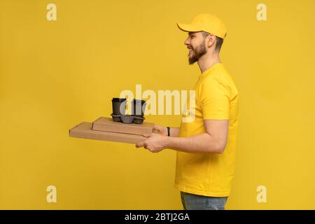 Portrait of young delivery man in yellow uniform with pizza boxes and takeaway coffee isolated over yellow background. Stock Photo