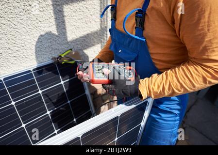 Man engineer installing solar photovoltaic panel system on roof of modern house. Electrician checking blue solar module, using voltmeter. Alternative energy ecological concept. Stock Photo