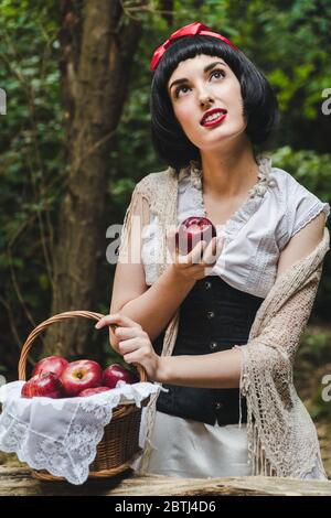 Snow White holds a red apple from a basket in her hand and looks up with a face full of dreams. Stock Photo