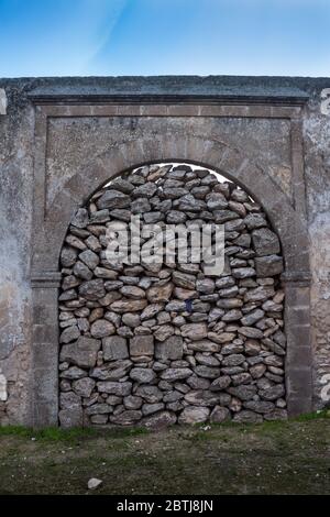 Fence with a gate with an arch, barred with stones. Part of an abandoned building. Blue sky with white clouds. Green grass. Dar Caid Hadji (Hajji), Mo Stock Photo
