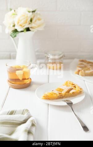 Piece of homemade shortbread lemon pie sprinkled with powdered sugar on a plate. Metal fork for a dessert, tea in a mug. White flowers in a jug. Stock Photo