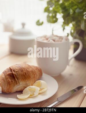 Fresh croissant with butter on a white plate and hot chocolate with melting marshmallow in a white cup on wooden table. Breakfast at home. Morning rel Stock Photo