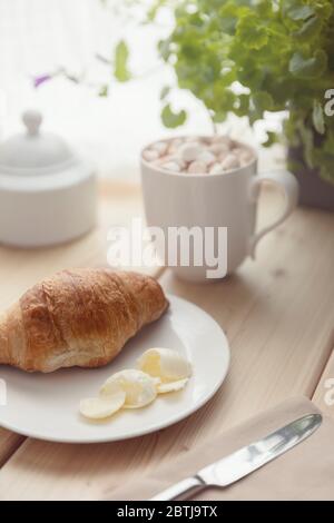 Fresh croissant with butter on a white plate and hot chocolate with melting marshmallow in a white cup on wooden table. Breakfast at home. Morning rel Stock Photo