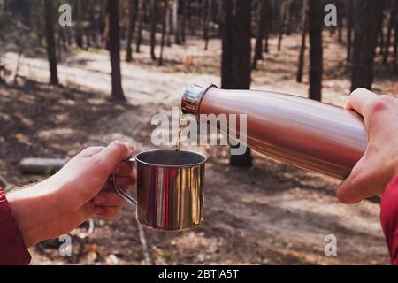 Man pouring tea from thermos in a metal cup while camping in pine forest. Stock Photo