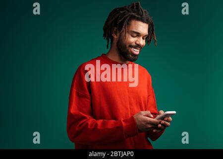 Portrait of a young african man wearing pullover standing isolated over green background, using mobile phone Stock Photo