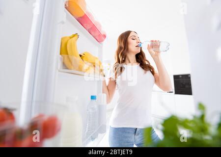 Portrait of charming lovely girl have morning running exercise want rest open fridge hold bottle drink water wear white t-shirt in modern kitchen Stock Photo