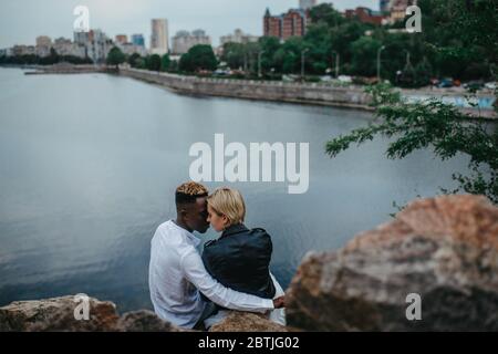 Interracial couple sits on rocks and hugs against background of river and city. Concept of love relationships and unity between different human races. Stock Photo