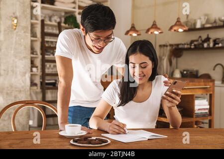 Image of multicultural smiling couple using cellphone and making notes in planner in cozy kitchen Stock Photo