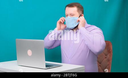 bearded man in a purple shirt is wearing a protective medical mask to work at a laptop at home on a green background. Quarantine concept Stock Photo