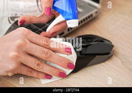 Woman cleaning black computer mouse, about to spray disinfect on it, holding back with paper tissue, closeup detail on hands with pink fingernails Stock Photo