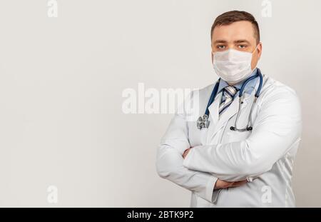 A male doctor in a medical mask with arms crossed, on his chest poses in front of the camera in a white coat near a white building during the COVID-19. Stock Photo