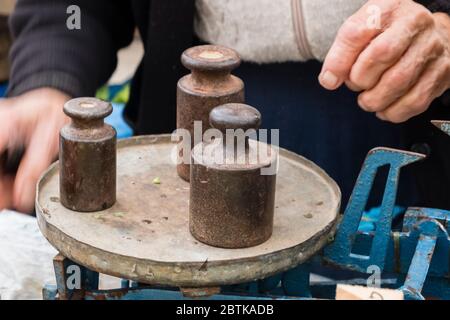 Older female street vendor uses an old mechanical scale with old metal weights to weigh fruits & vegetables for sale, at Split's Green Market, Croatia