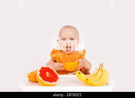 Happy baby sitting in high chair eating fruits in a white kitchen. Healthy nutrition for kids. Children eat oranges and grapefruits. Stock Photo