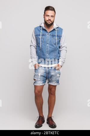 Stylish young cheerful guy with a beautiful beard in a denim jacket, a white T-shirt and denim shorts on a simple gray background. Denim jacket and Stock Photo