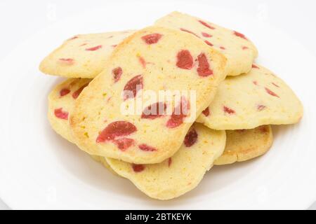 A pile of traditional, crispy slice and bake cookies, home-baked golden biscuits with pieces of red cherry presented on a white plate Stock Photo