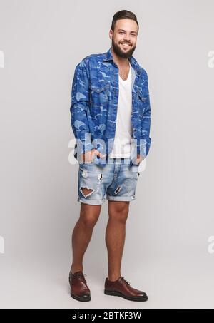 Stylish young cheerful guy with a beautiful beard in a denim shirt and denim shorts on a simple gray background. Denim shirt and denim shorts Stock Photo