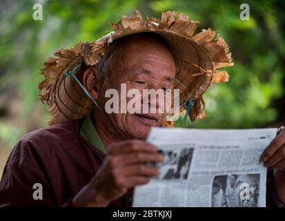 HOI AN - MAR 10, 2018: An old man with an Asian conical hat reading newspaper outdoor under a street on the street in Hoi An, Vietnam on 10 March 2018 Stock Photo