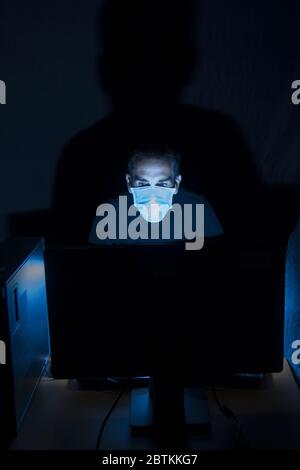 an adult man wearing protective mask and using computer in a dark room ,computer screen is the only source of light projecting a big shadow on backgro Stock Photo