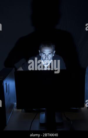 an adult man wearing protective mask using computer in a dark room ,computer screen is the only source of light projecting a big shadow on background Stock Photo