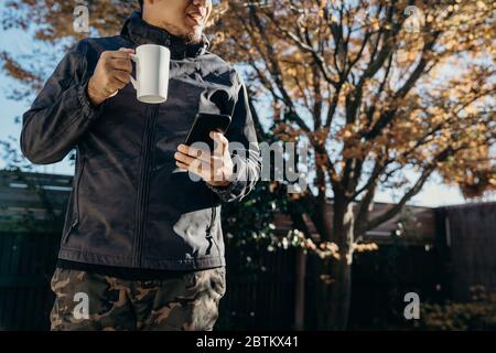 Asian man Standing at backyard using mobile phone and drinking coffee. Man relaxing at home reading text message on his cell phone in morning. Stock Photo