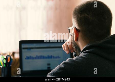 shot from back of pensive businessman with glasses looking at the screen of a laptop. Pensive confused young man thinking of problem solution stuck wi Stock Photo