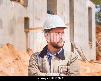 Caucasian bearded civil engineer or construction worker in white hardhat stands cross handed opposite construction site. Stock Photo