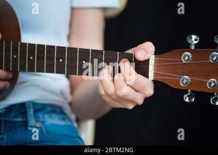 Close-up of girl's hand playing G major chord on tenor ukulele while seated, with black background and copy space Stock Photo