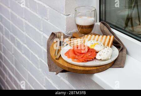 Toasted bread, salted salmon and poached egg with cream cheese and cup of latte macciato on white windowsill. Stock Photo