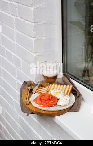 Toasted bread, salted salmon and poached egg with cream cheese and cup of latte macciato on white windowsill. Stock Photo