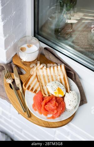 Toasted bread, salted salmon and poached egg with cream cheese and cup of latte macciato on white windowsill. Stock Photo