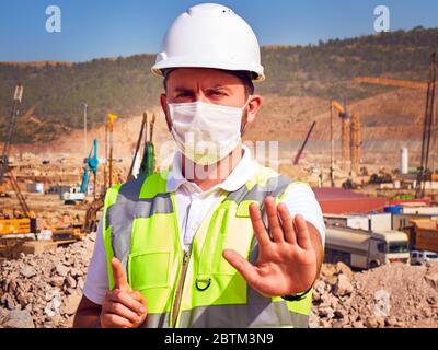 Engineer in medical mask shows stop sign standing opposite construction site. Stock Photo