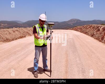 Geodetic engineer surveyor in white hard hat doing measurements with GNSS satellite receiver during road construction works. Stock Photo