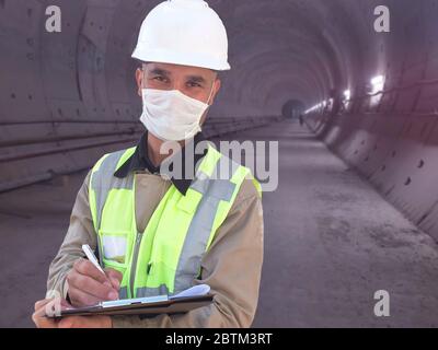 Construction worker dressed in medical mask, uniform and white hardhat stands with clipboard at metro tunnel under construction Stock Photo