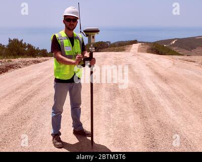 Geodetic engineer surveyor in white hard hat doing measurements with GNSS satellite receiver during road construction works. Stock Photo